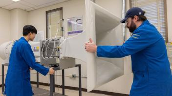 Two individuals in blue lab coats maneuvering large sound equipment in a lab setting.