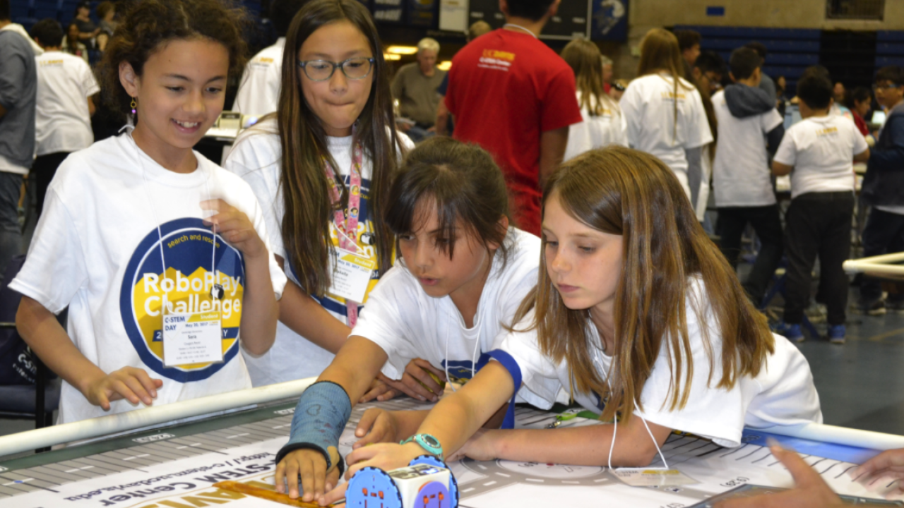 Young students wear white T Shirts with event logo and work on robotics projects indoors