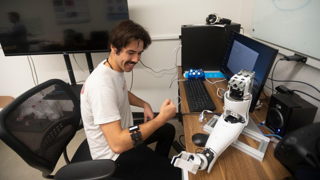 Person sits at a desk wearing a mechanical arm band and looking at a robotic arm