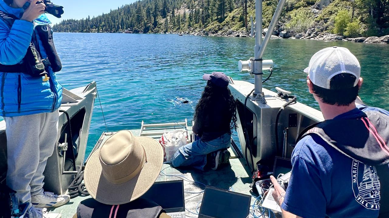 The student team on a small boat looking toward Lake Tahoe and lush green trees on the shoreline