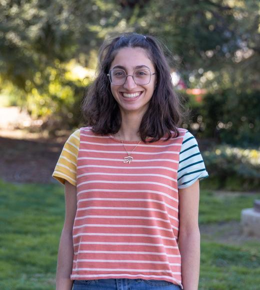 Portrait of Anna Rita Moukarzel standing outside wearing a colorful striped shirt with greenery in background
