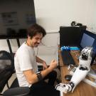 Person sits at a desk wearing a mechanical arm band and looking at a robotic arm