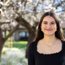 A young woman with long brown hair wearing a black long-sleeved shirt stands in front of blossoming trees